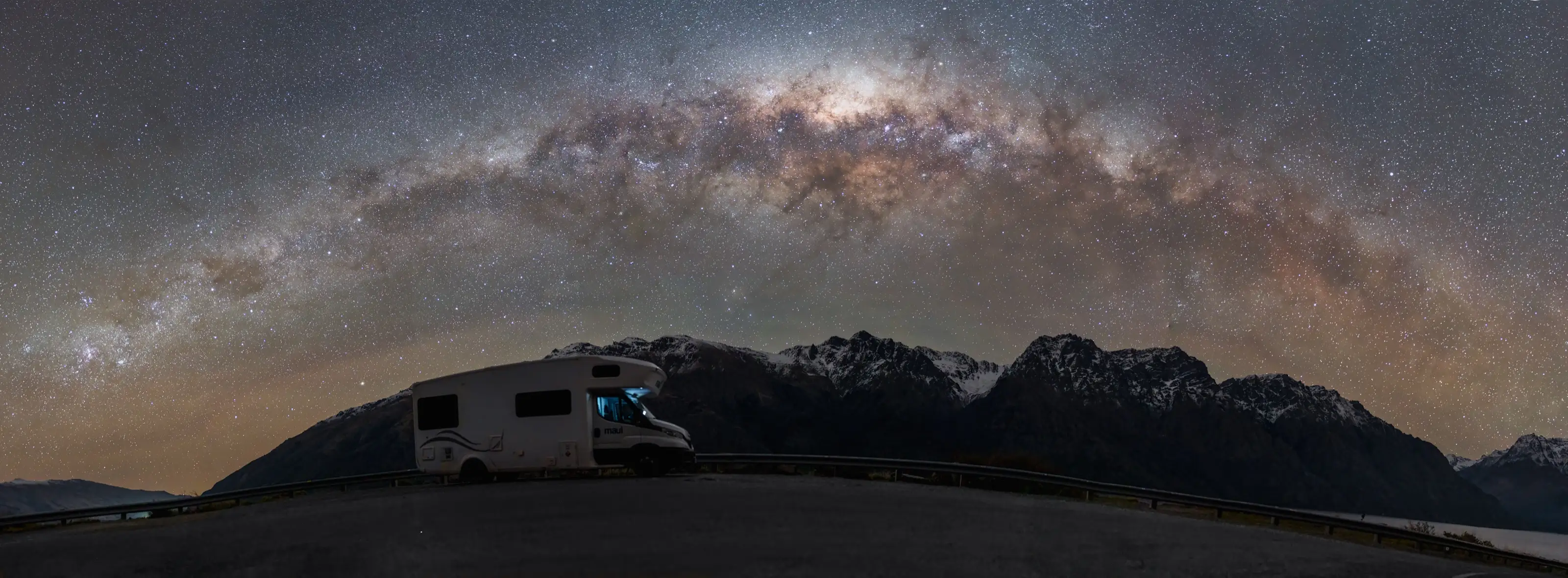 The Milky Way arching over a snow-capped ridge, a campervan parked below